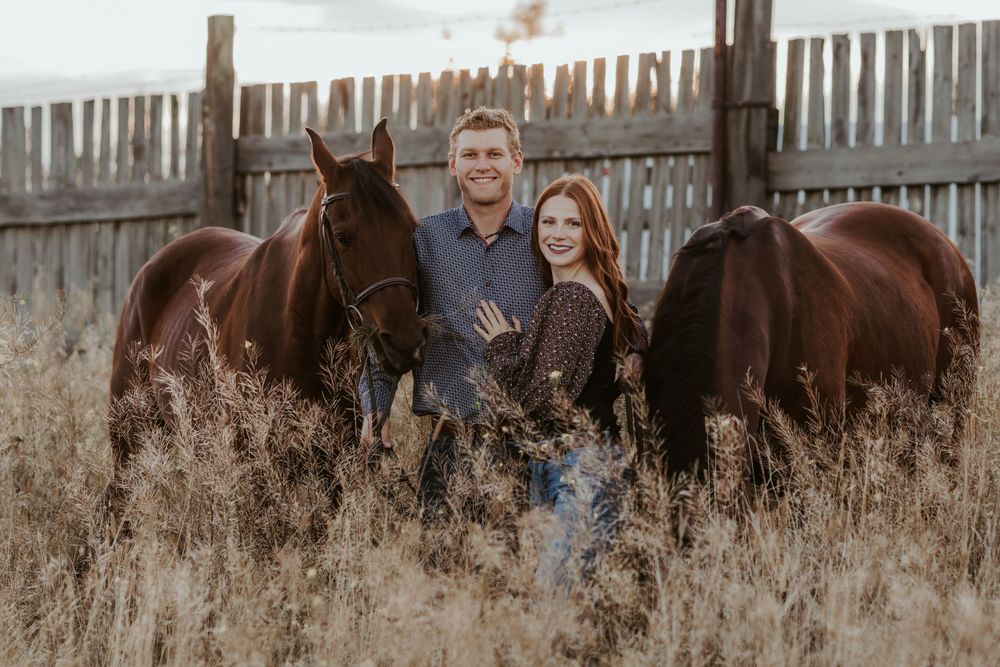 Smiling couple standing together between two dark horses near a rustic wooden fence.