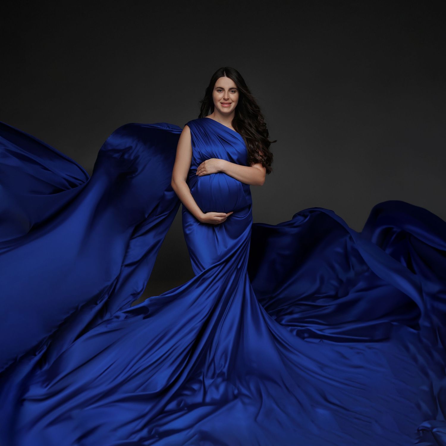 Flowing royal blue silk maternity gown with dramatic movement captured against dark background in elegant portrait.