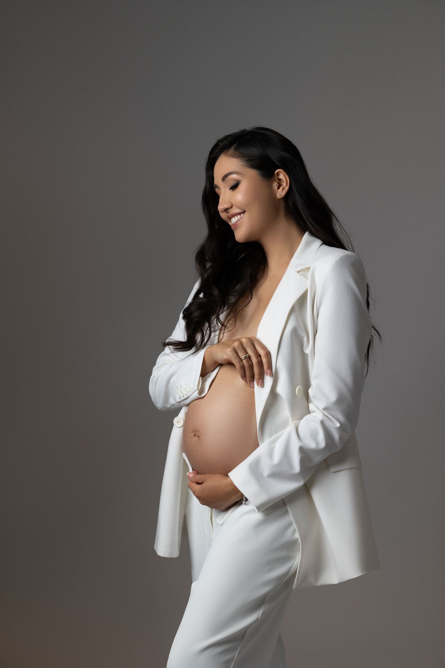 A pregnant woman in a white outfit poses gracefully against a gray studio backdrop for an artistic maternity photoshoot.