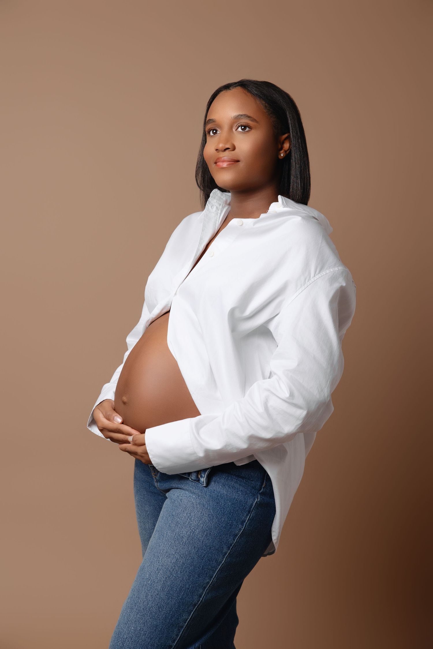 Casual maternity portrait with white button-down shirt and jeans against neutral background.