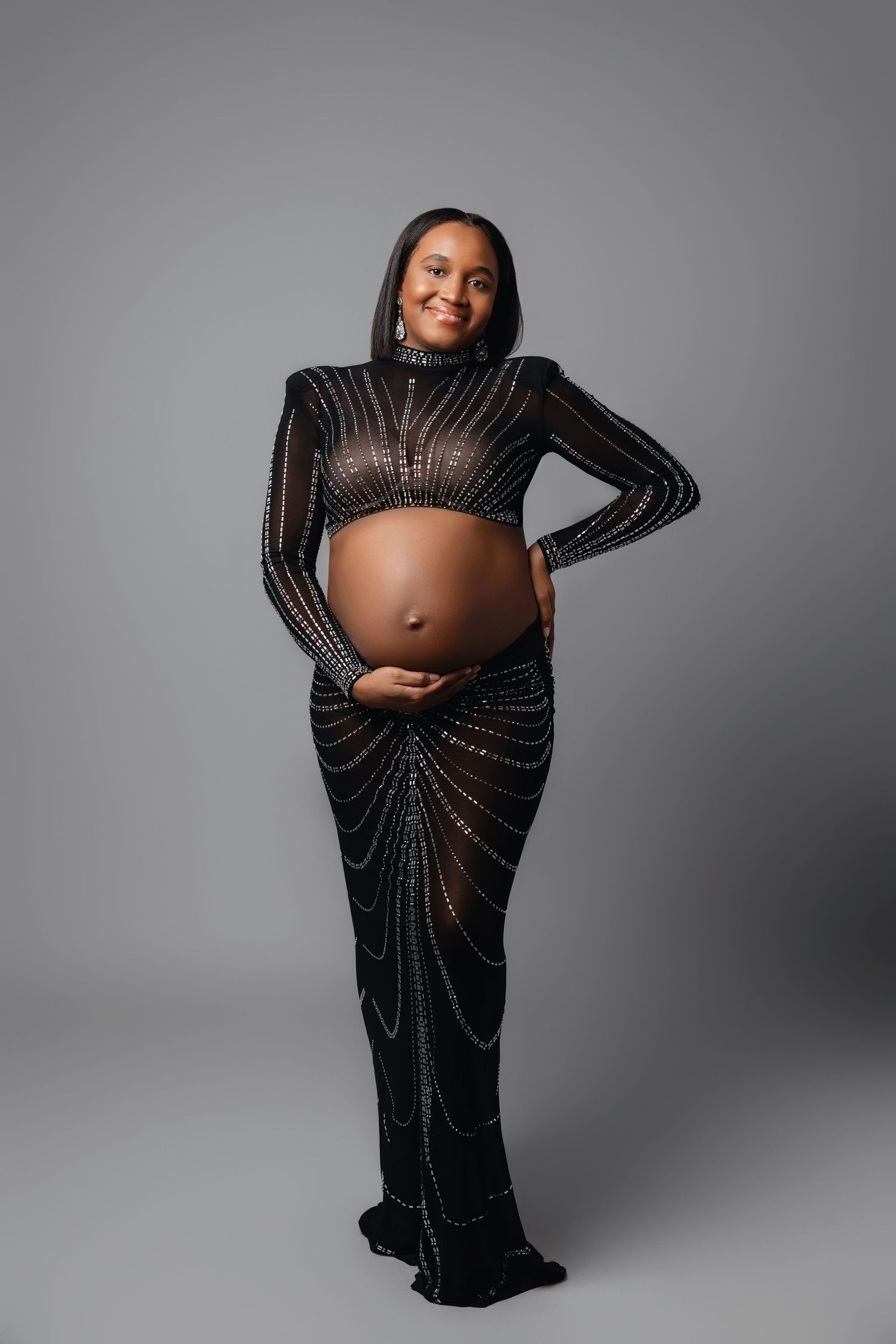 A pregnant woman poses in a glamorous black beaded gown with long sleeves showing her baby bump against a gray backdrop.