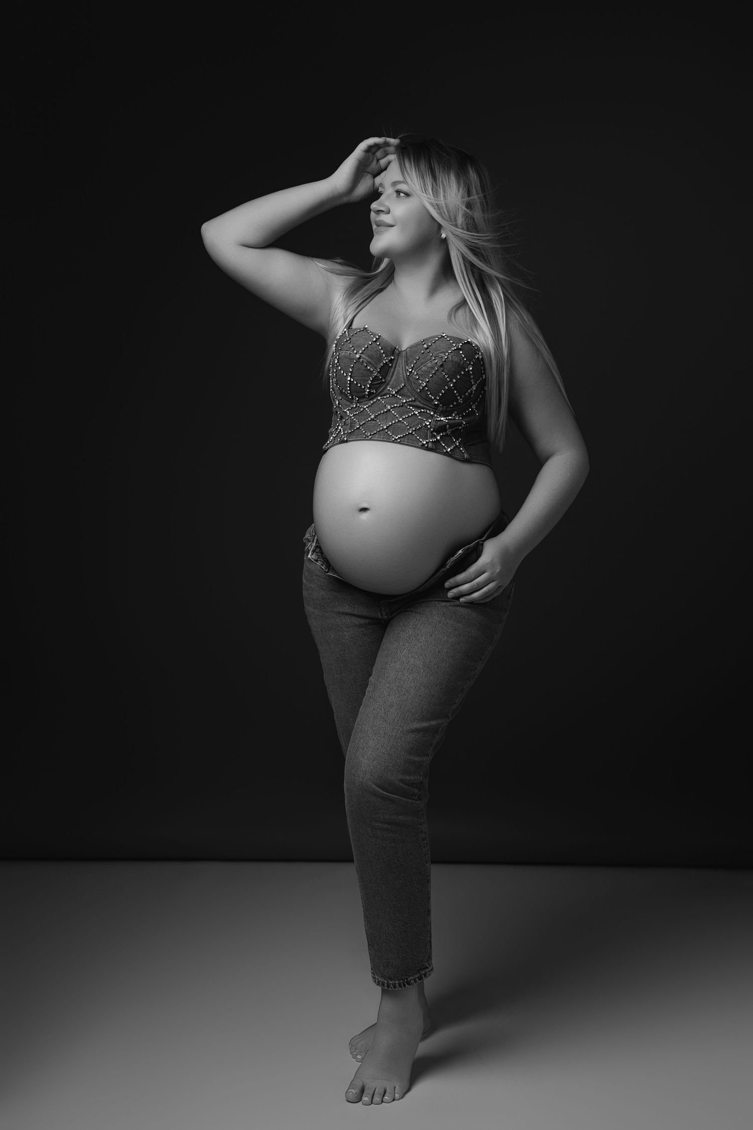 Black and white maternity photo of expectant mother posing in jeans and patterned top against dark background.