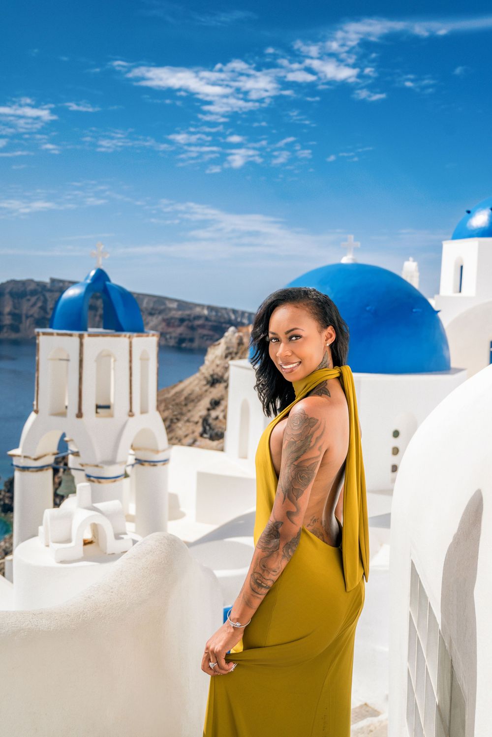A person in a yellow dress poses against the iconic white buildings and blue domes of Santorini, Greece under bright skies.