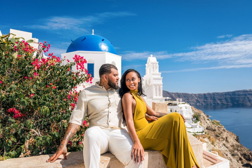 Couple enjoying scenic views of white buildings and blue sea in Santorini, Greece with vibrant bougainvillea flowers.
