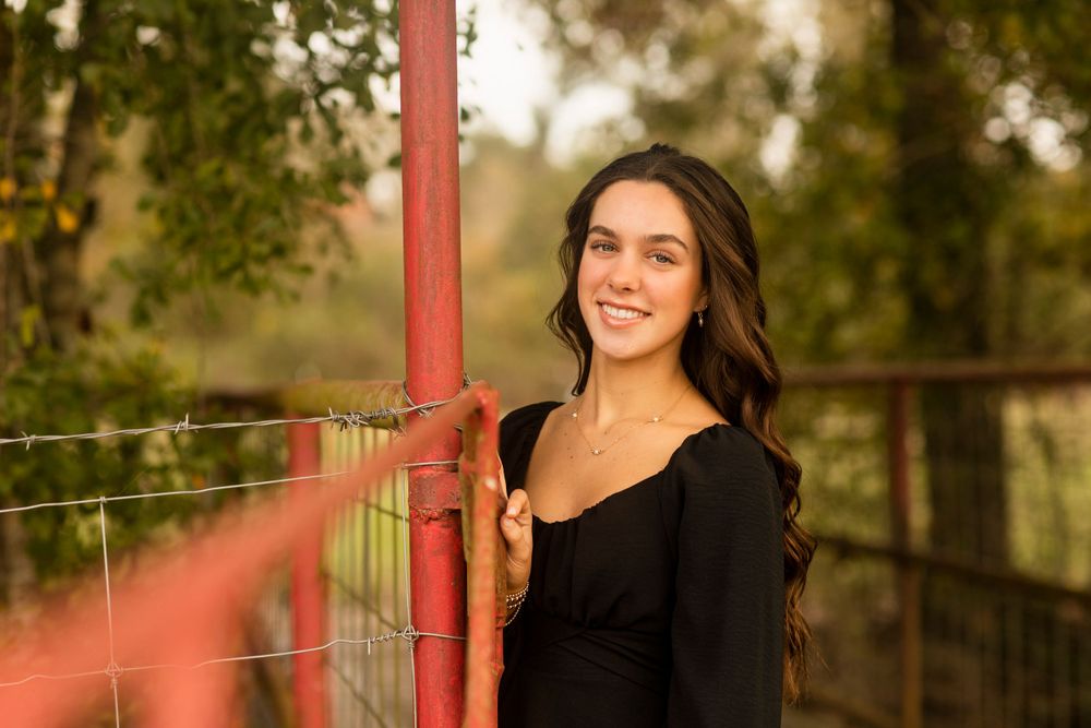 Senior graduate portrait by a rustic red gate and fence in warm sunset lighting.