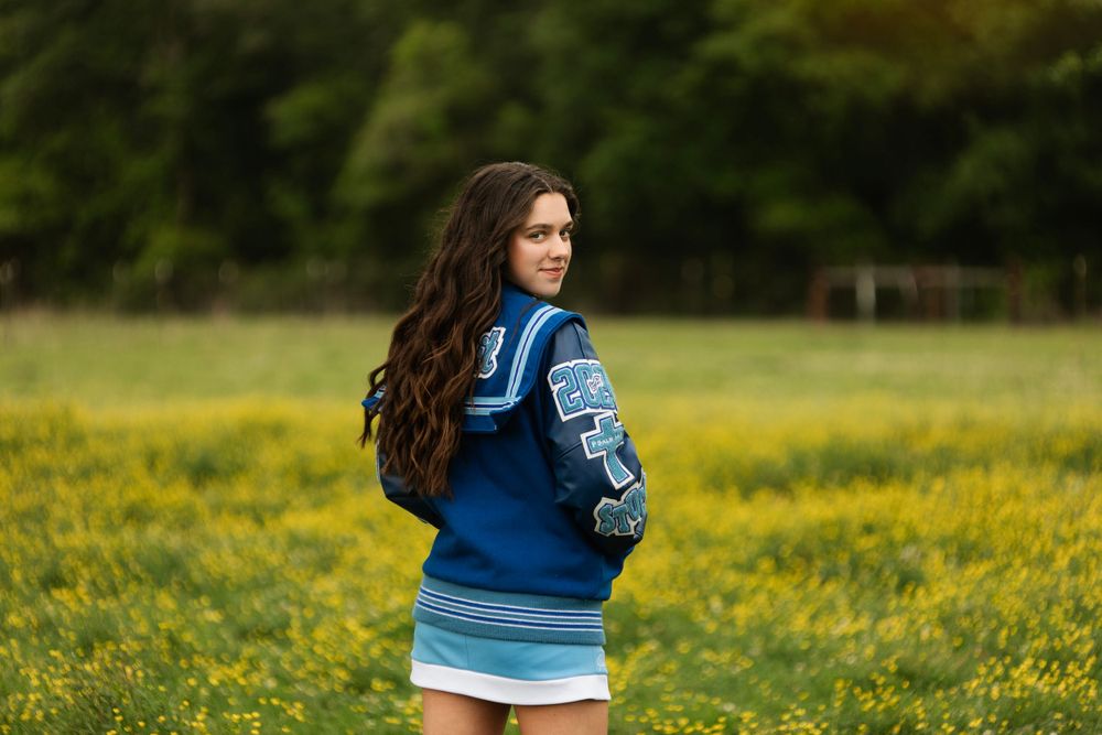Senior graduate portrait with blue varsity jacket against backdrop of yellow wildflowers in field.