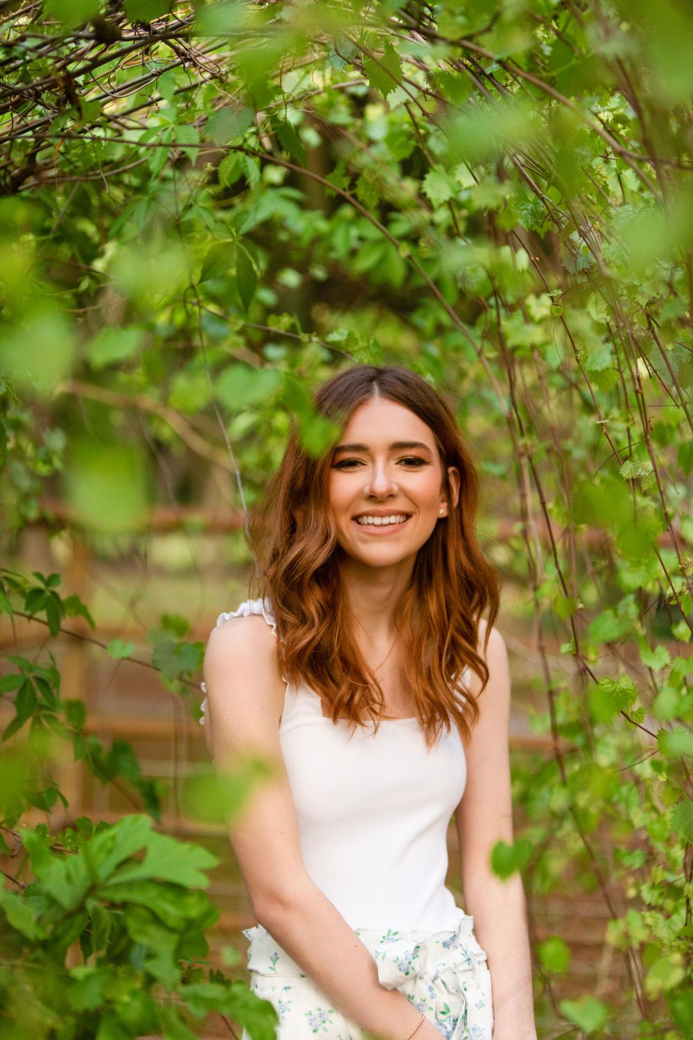 Spring senior graduate portrait among green foliage wearing white top.
