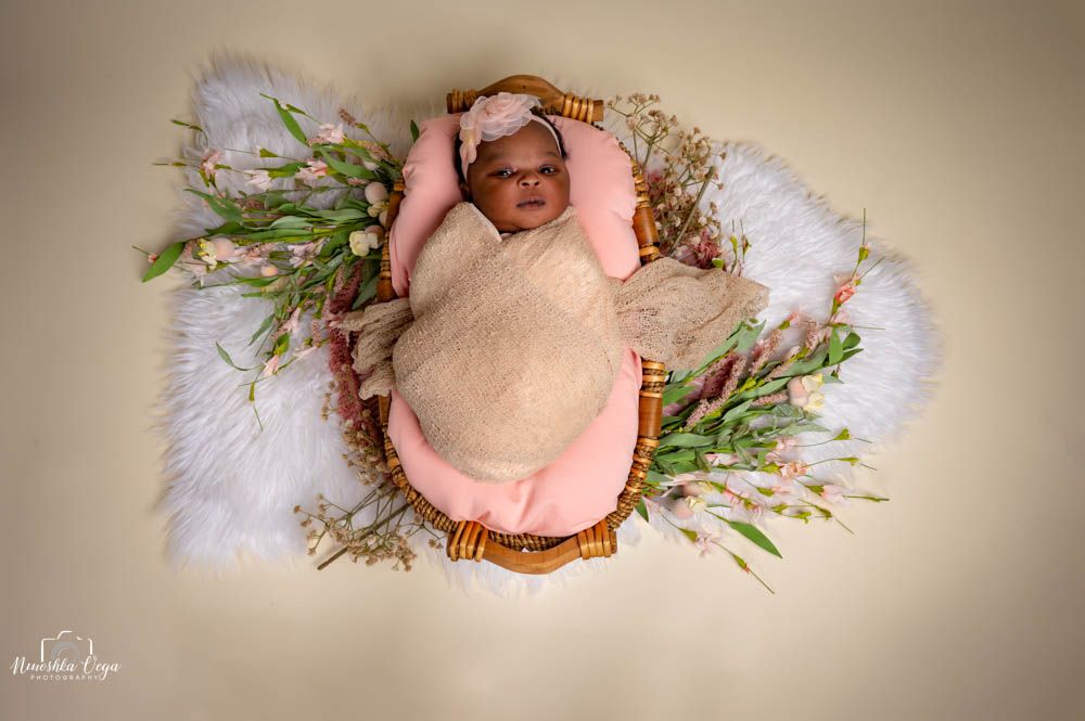 Newborn baby wrapped in pink blanket rests on cream backdrop with floral decorations.