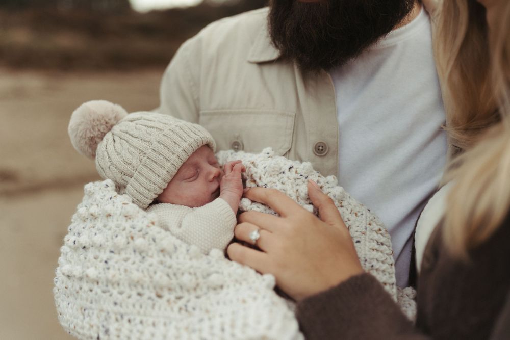 A sleeping newborn baby wrapped in a cozy white knit blanket wears a matching beanie hat while being cradled.