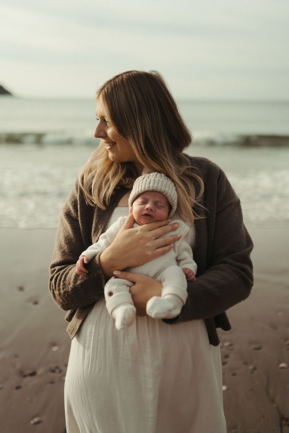 A parent in a brown sweater holds a newborn baby in white clothing while standing on a misty beach.
