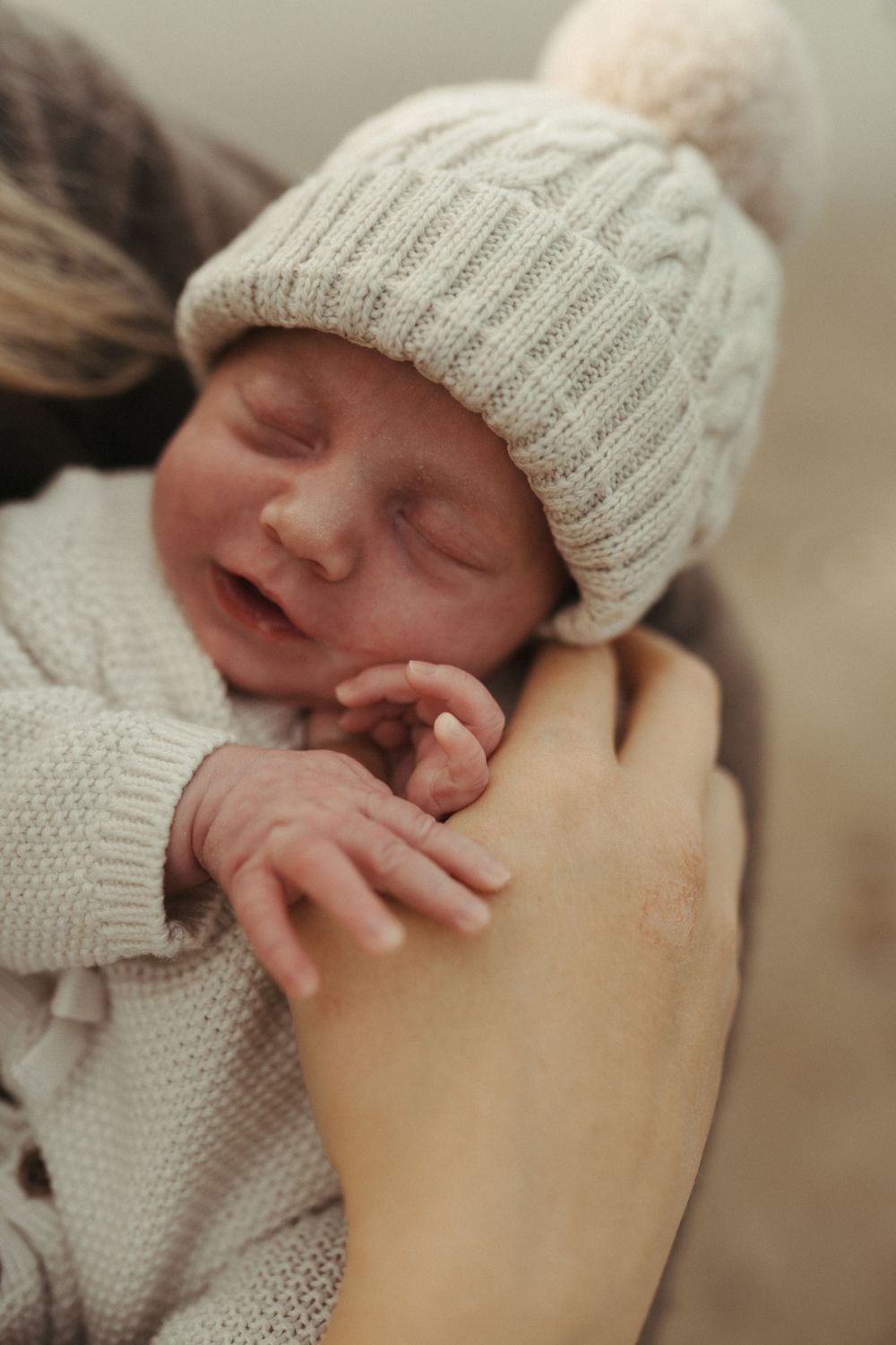 A sleeping baby wearing a cozy knit beanie and sweater is cradled in someone's arms.