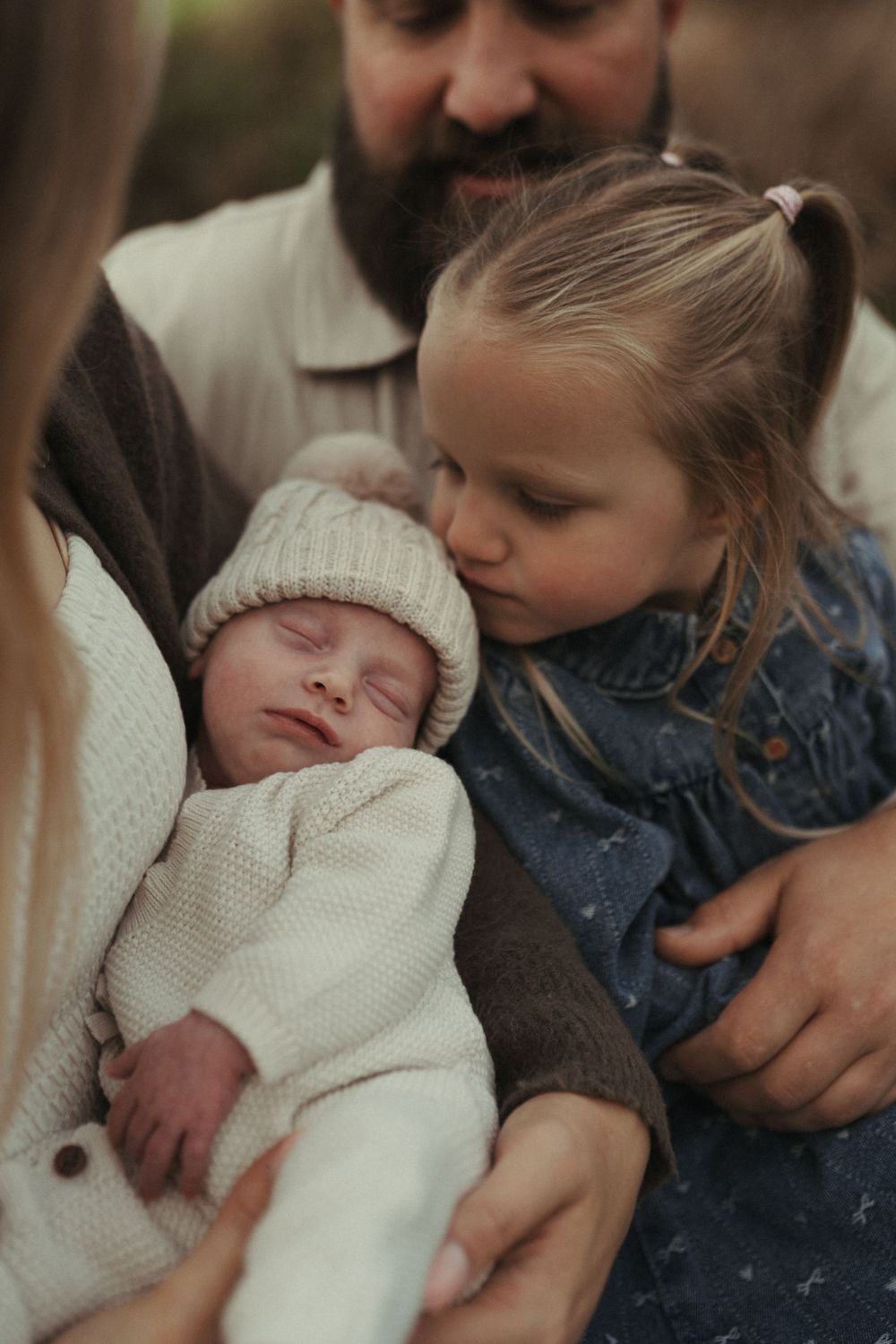 A tender family moment shows a newborn baby in a white knit hat being cradled while an older sibling looks on.