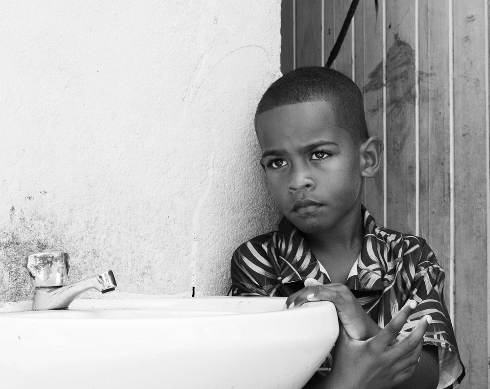 A black and white portrait photograph showing a young person resting their arm near a sink.