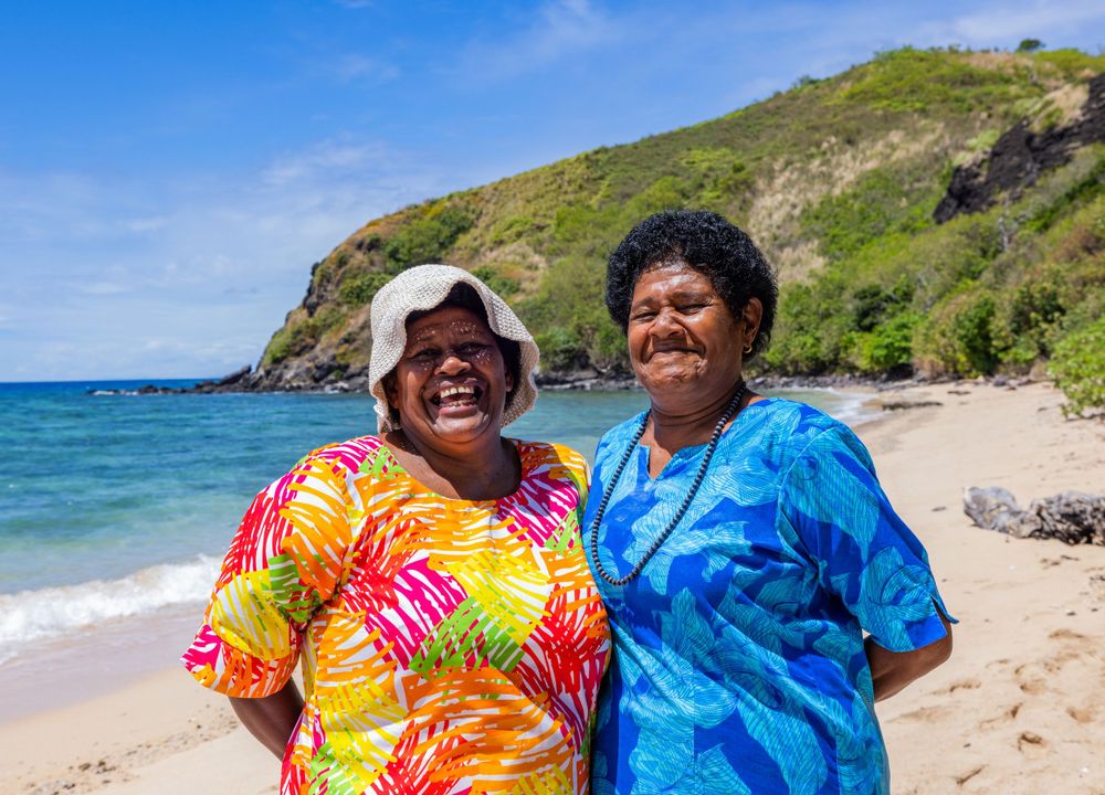 Two women in colorful shirts enjoying a sunny day at a tropical beach with hills in the background.