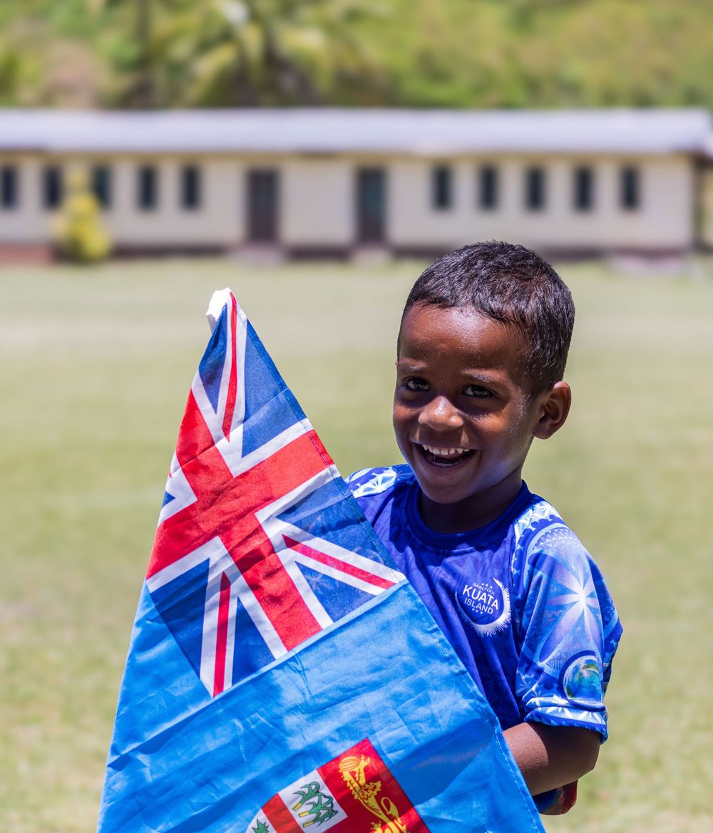 A cheerful youngster holds up a Fijian flag while wearing a blue shirt outdoors.