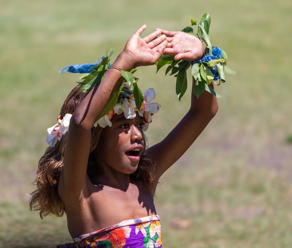 A person wearing floral decorations dances with raised arms in a grassy field.