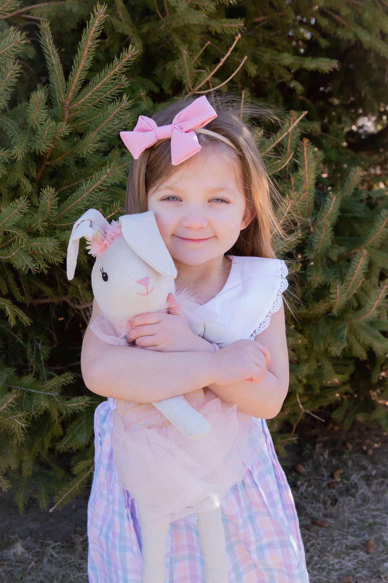 Child in pastel dress hugs a white plush bunny toy near evergreen trees.