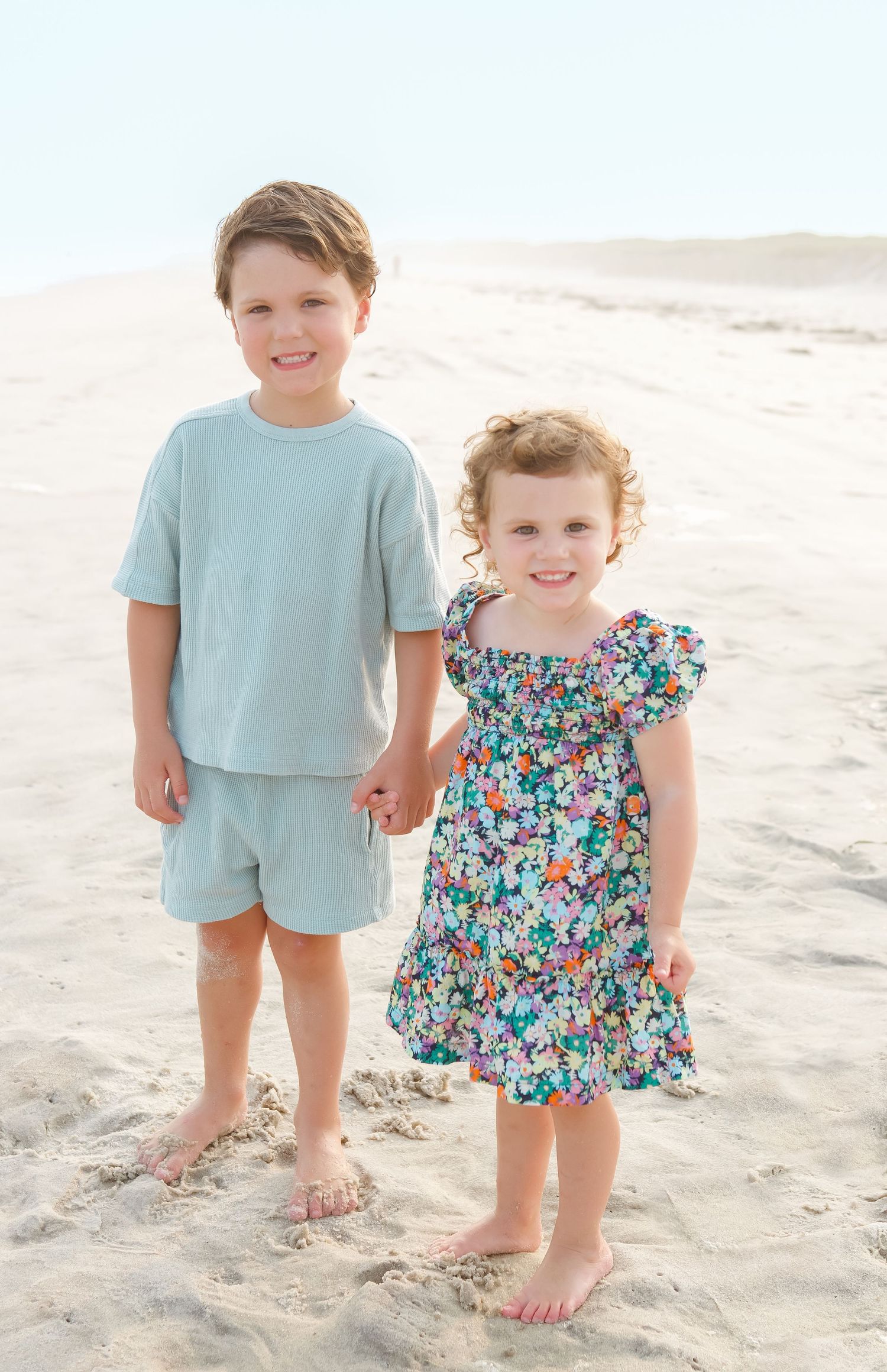 Two children in light blue and floral outfits play together on a sandy beach.