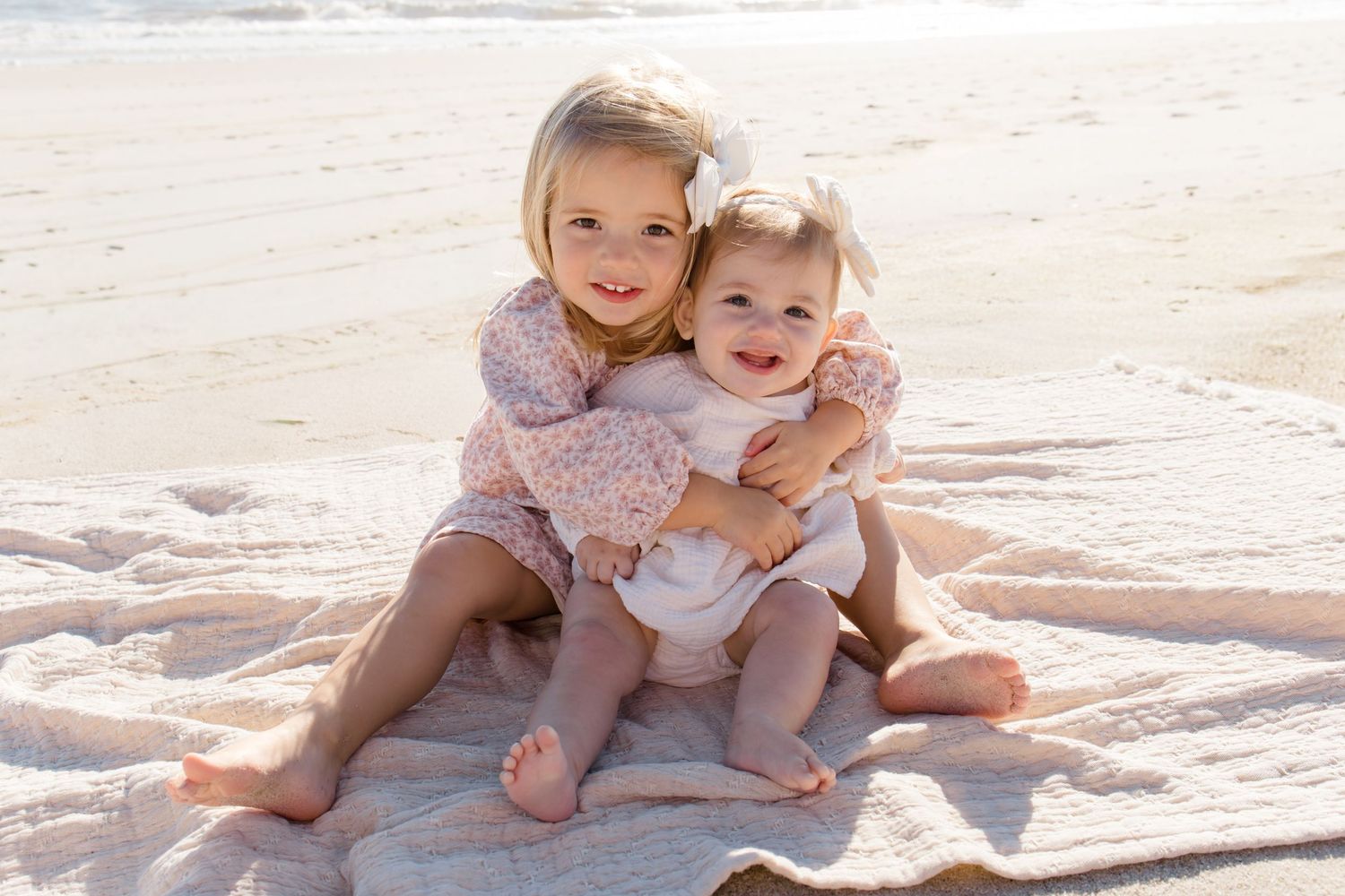 Two young siblings in floral dresses sitting together on a blanket at the beach during sunset.