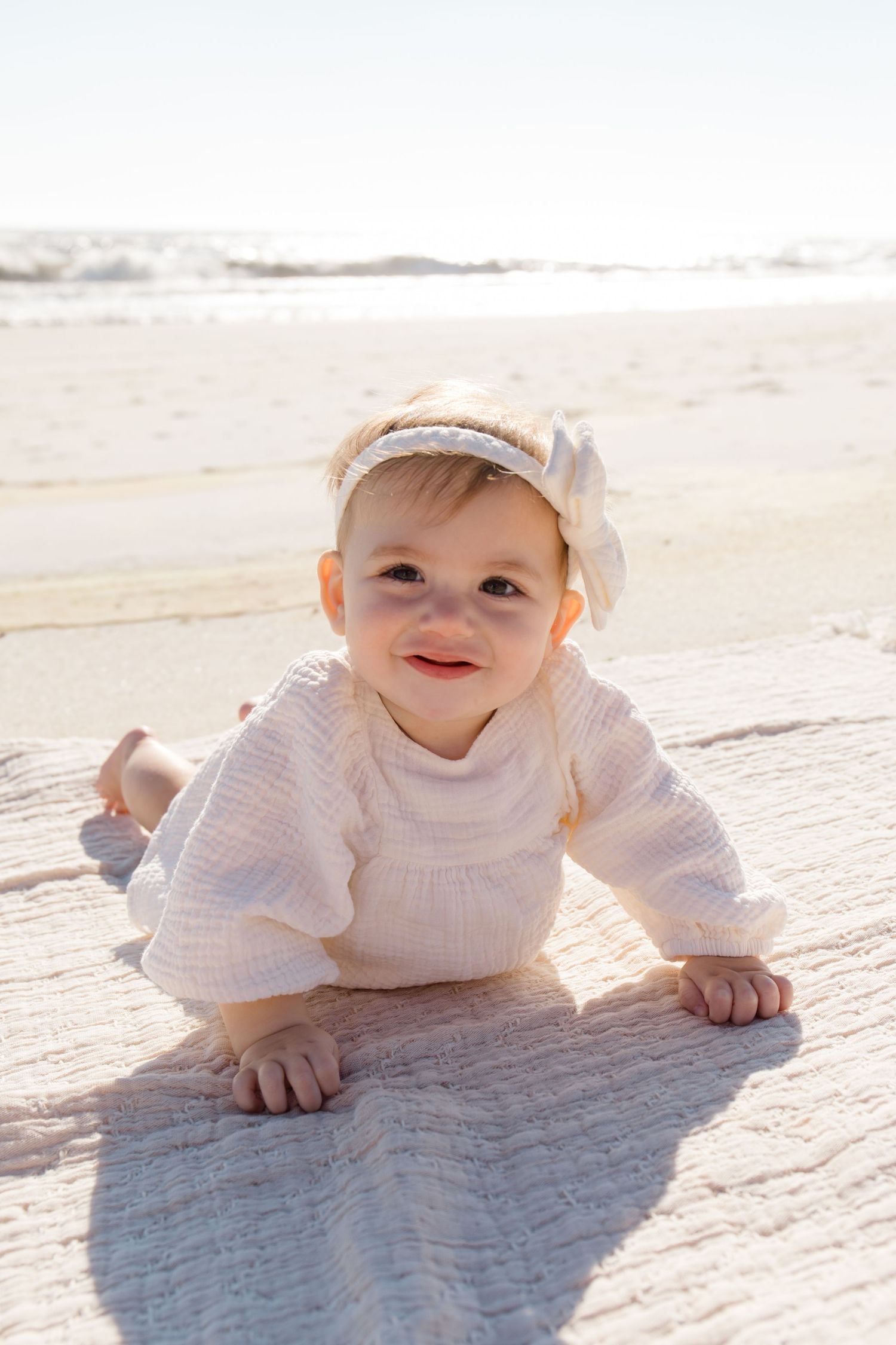 Baby wearing white sweater and cap crawling on blanket at sunny beach.