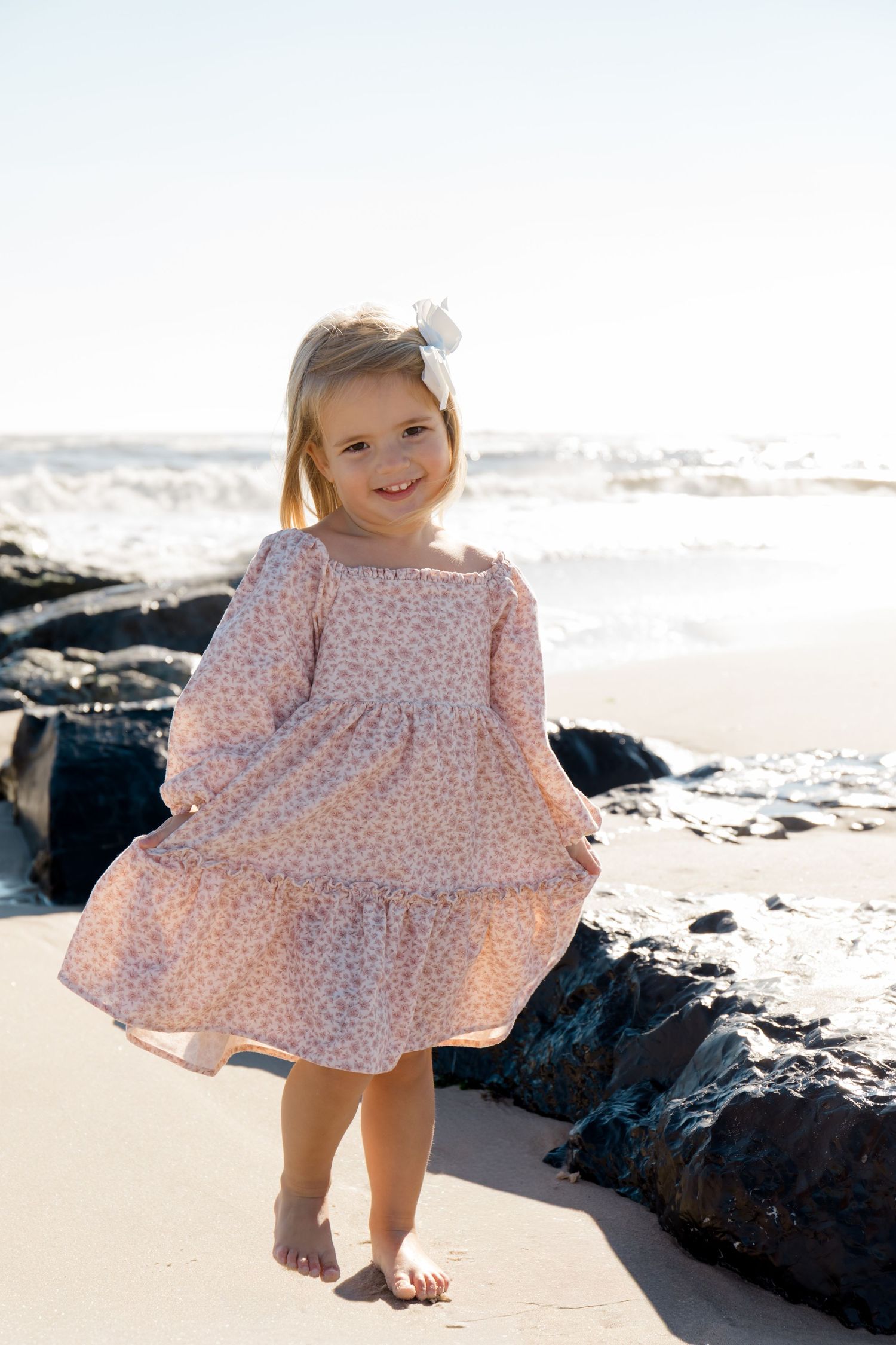 Young child in pink floral dress twirling on rocky beach at sunset.