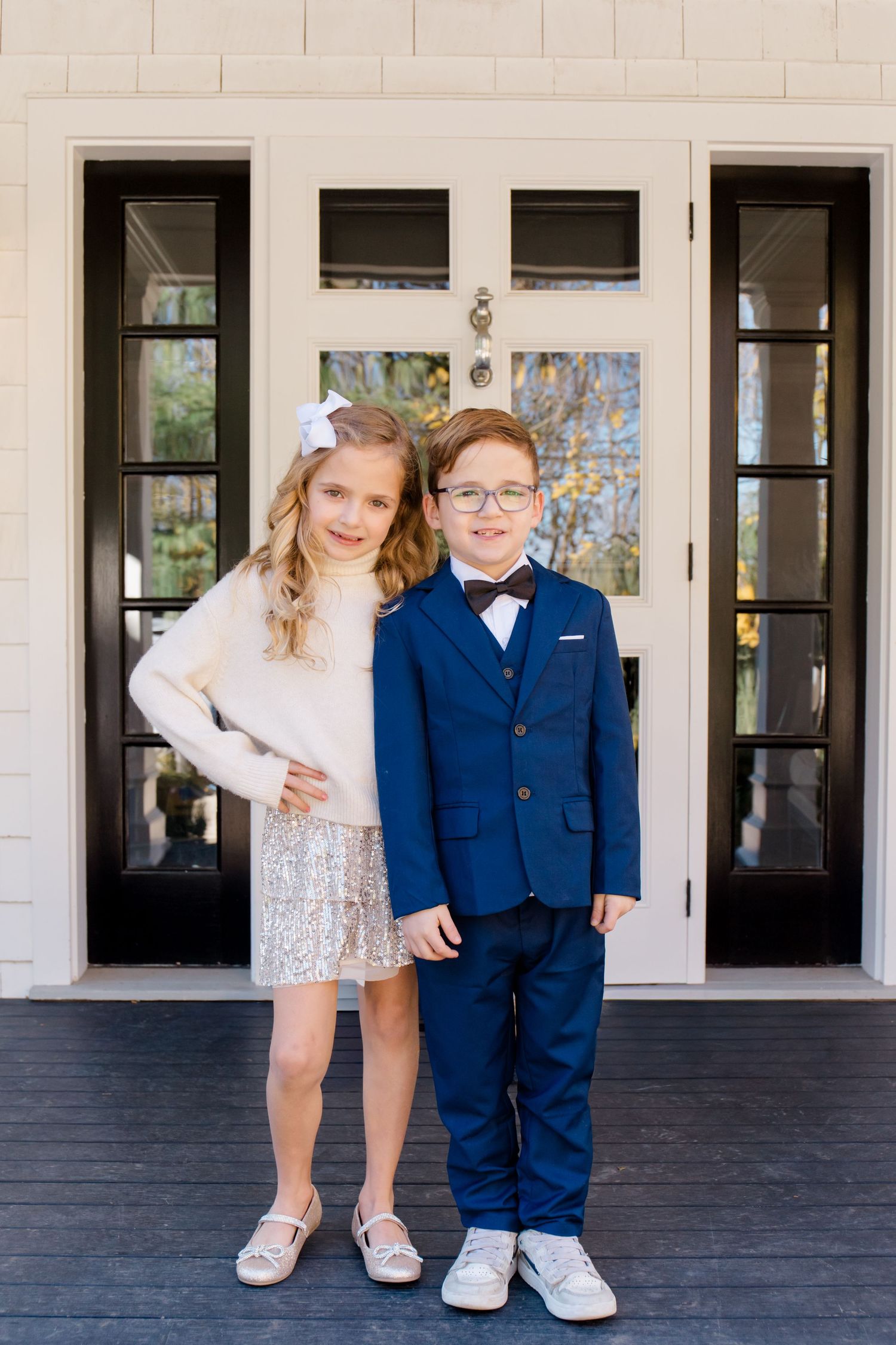 Two young children in formal attire stand together on a porch with white columns and dark wood flooring.