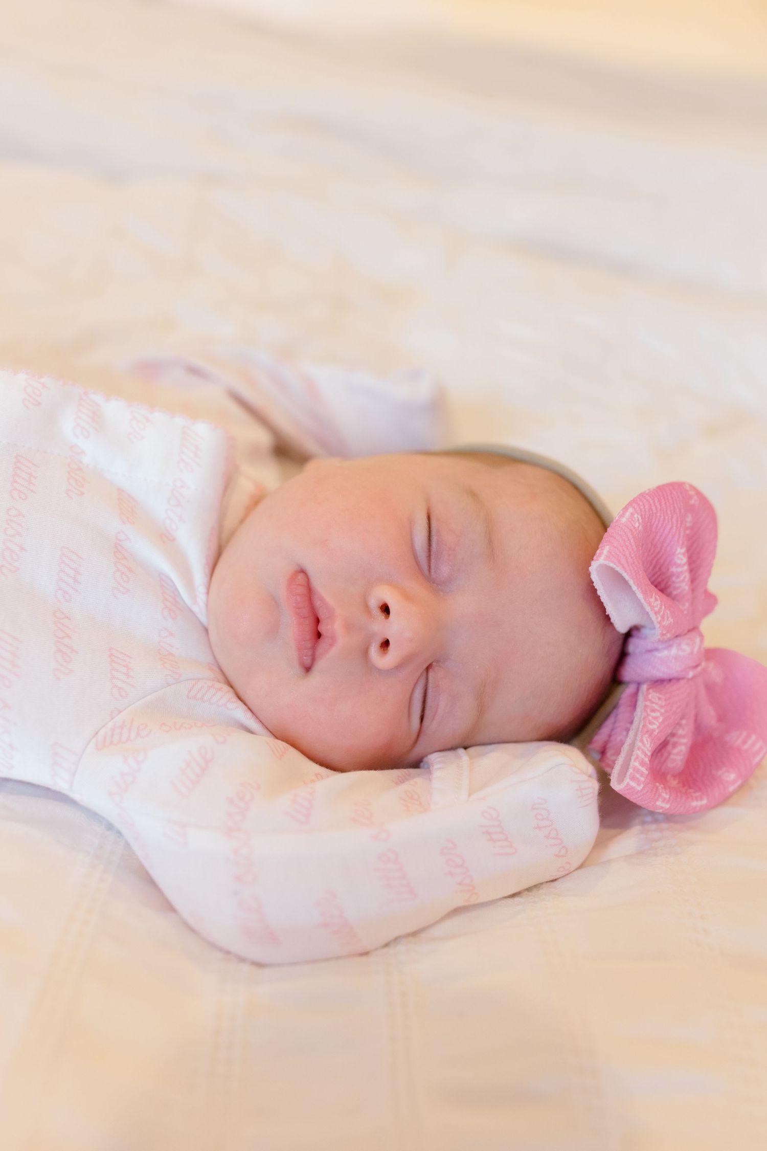 A sleeping newborn baby wearing a pink bow headband rests peacefully on cream-colored bedding.