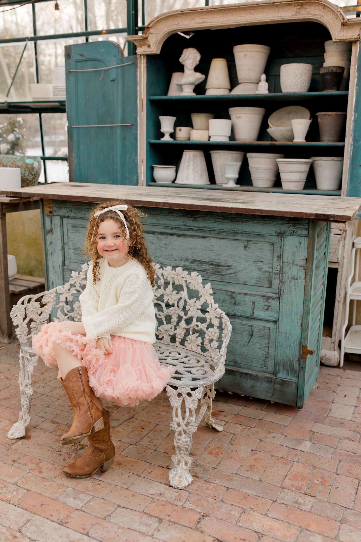 A vintage teal hutch with ornate white iron bench displays pottery on brick patio in rustic outdoor setting.