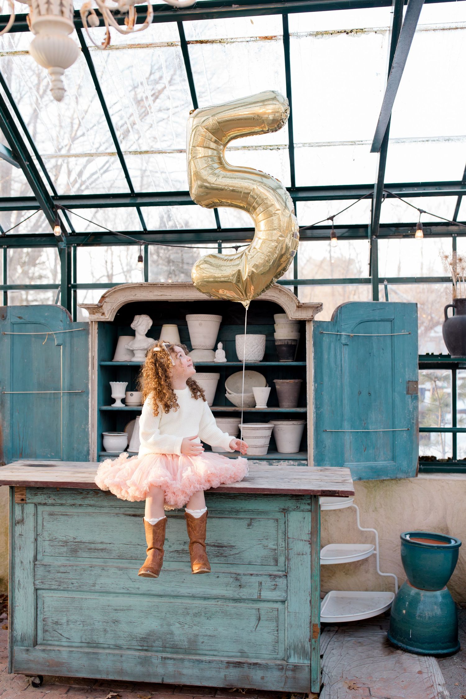 A child in a tutu sits on a vintage turquoise cabinet under a gold number 5 balloon in a greenhouse setting.