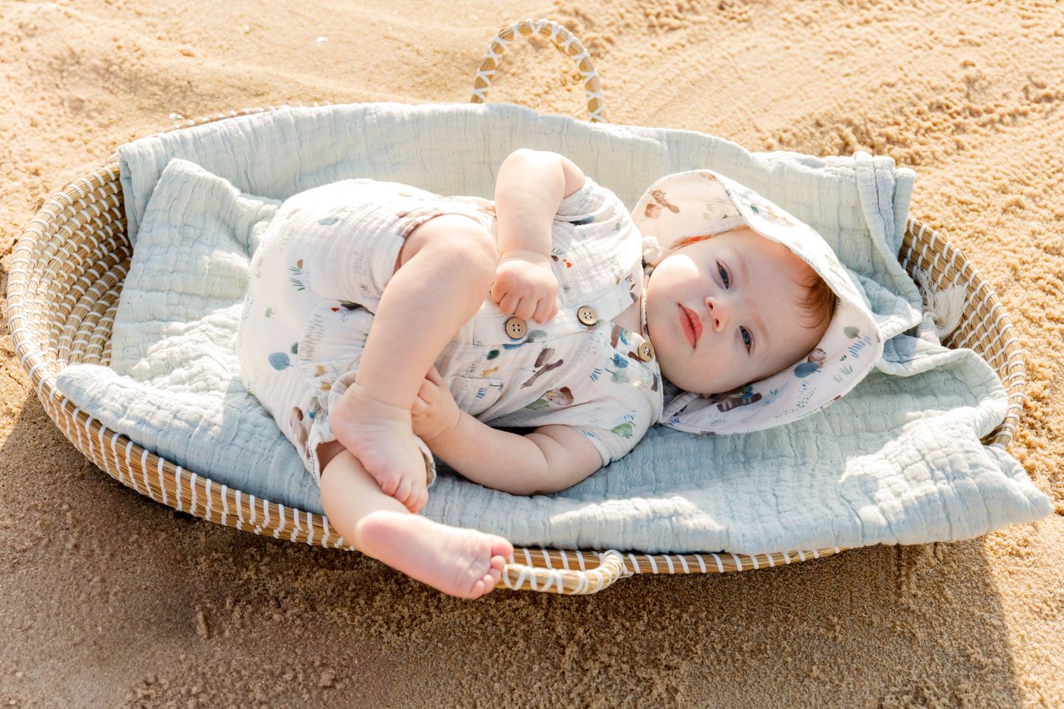 A baby rests peacefully in a woven basket on a sandy beach during a sunny day.