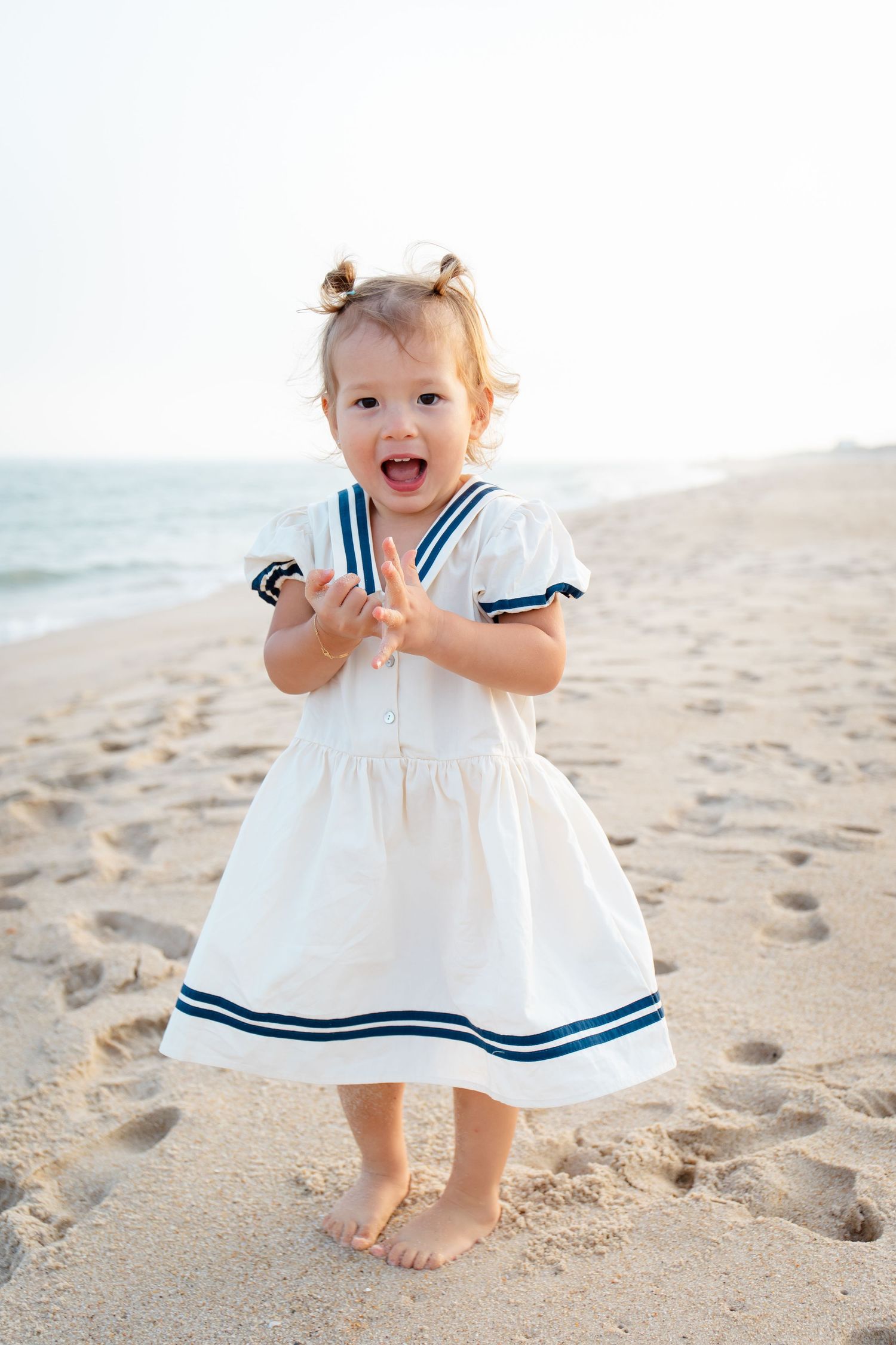 Toddler in white nautical dress playing on sandy beach.