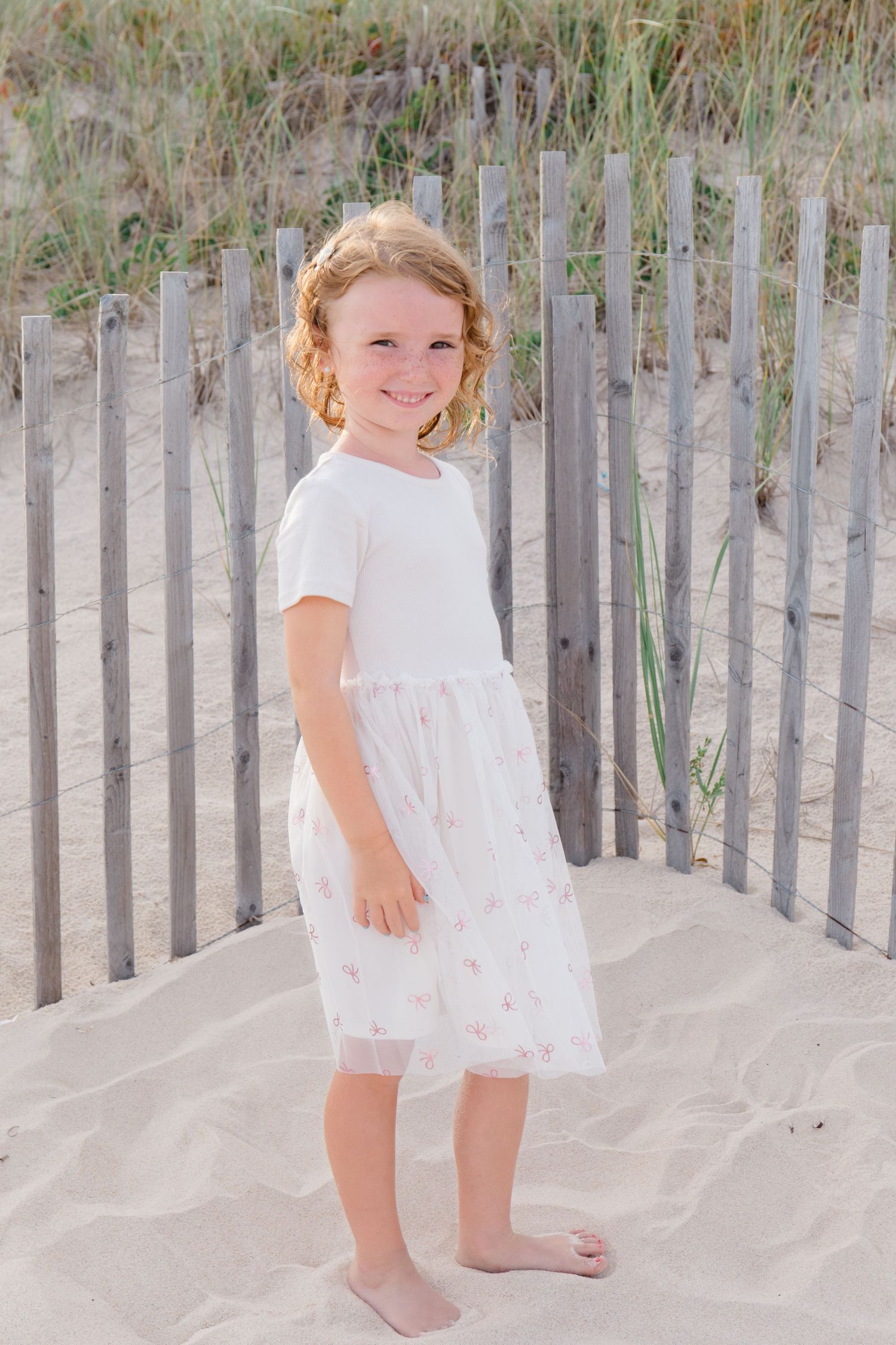 Young child in white dress exploring sandy beach area with wooden fence posts.