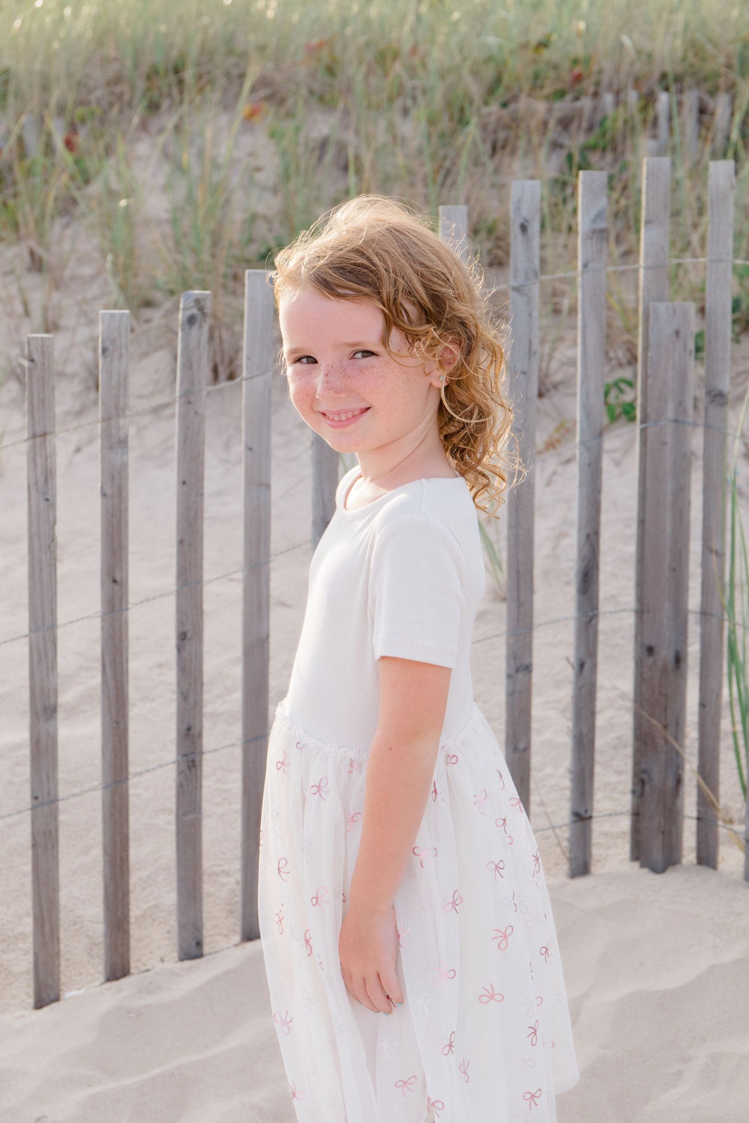 Child in a white dress walking along a sandy beach path with wooden fence posts at sunset.