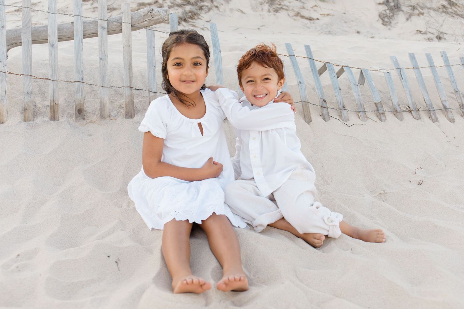 Two children in white dresses sitting together on sandy beach with fence in background.