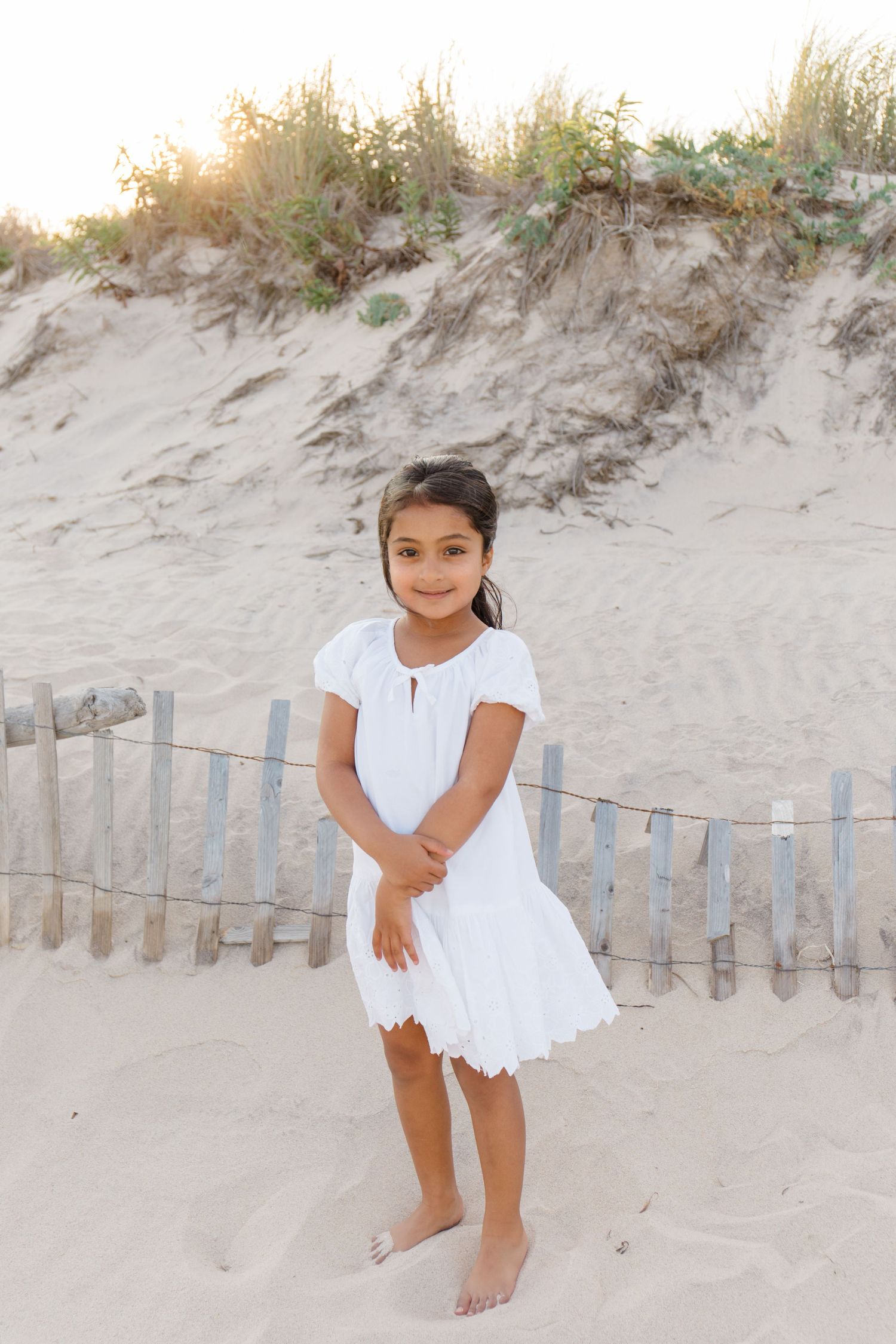 Child in white eyelet dress standing barefoot on sandy dunes with wooden fence behind.
