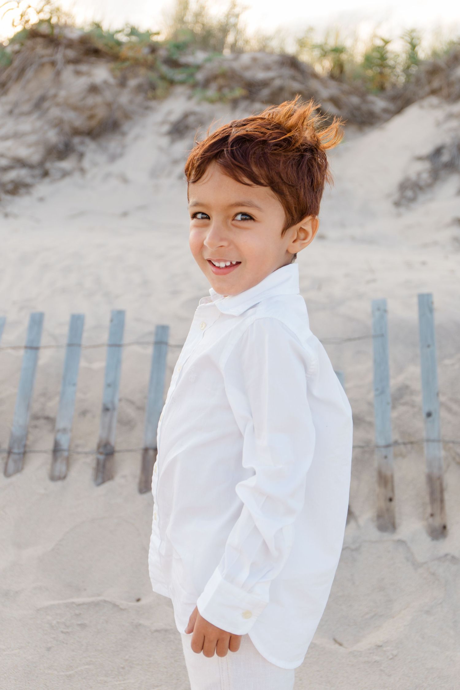 Child in all white outfit smiling against sandy beach dunes and wooden fence background.
