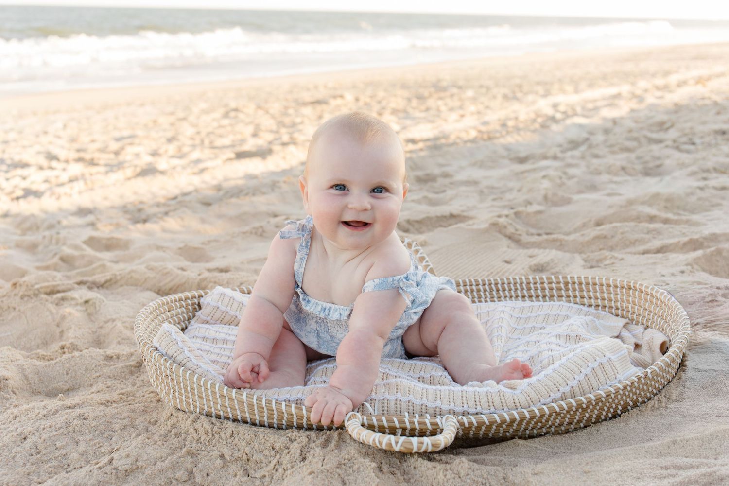 A baby lies on a round woven mat on a sandy beach during a sunny day.
