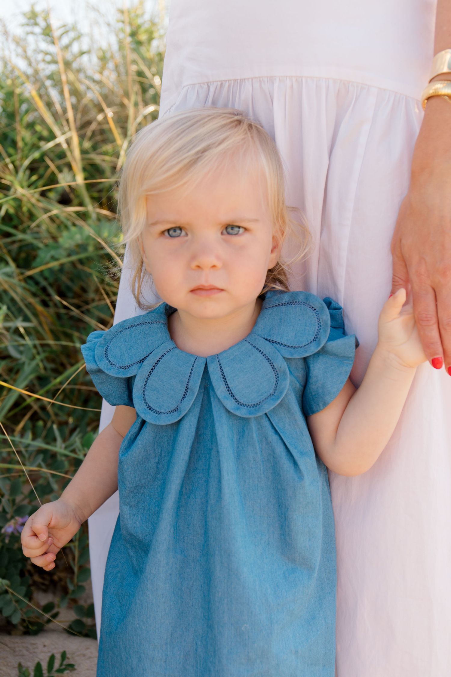 A toddler wearing a blue dress with a scalloped collar stands in tall grass while holding onto someone's white dress.