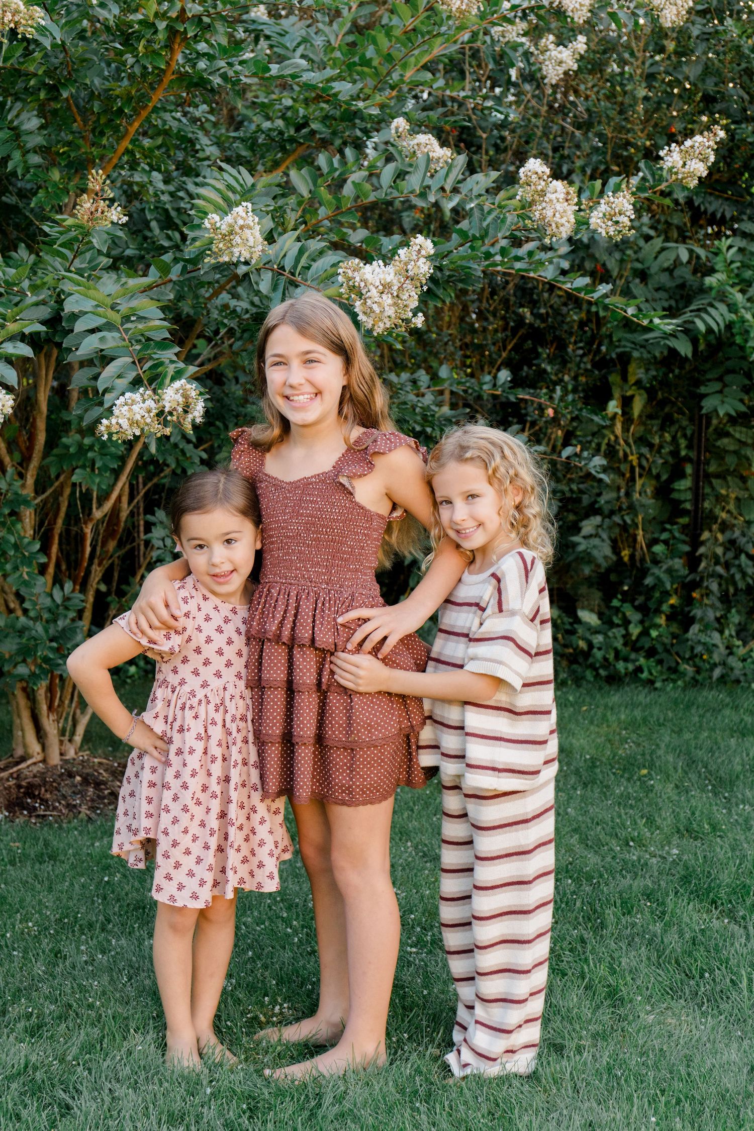 A group poses together in a garden setting wearing casual summer outfits against lush green foliage.