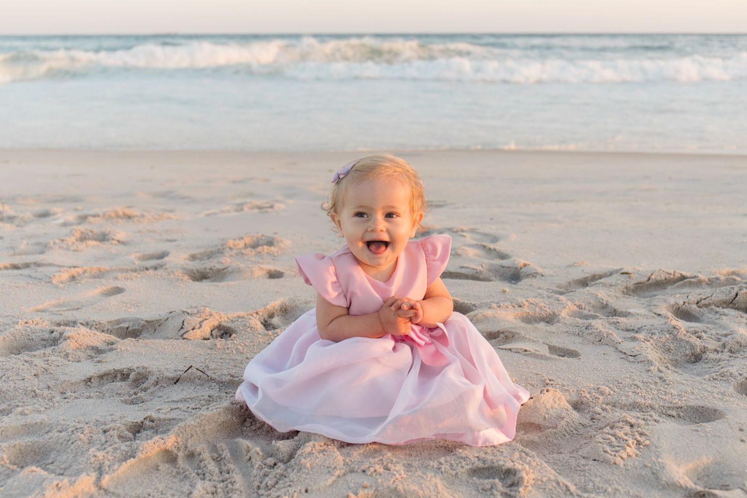 Baby in pink dress sitting and laughing on sandy beach with ocean waves behind.