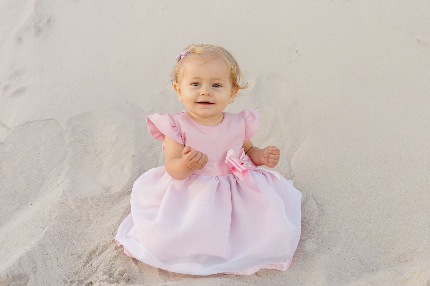 Baby wearing pink dress sitting on sandy beach during sunset photoshoot.