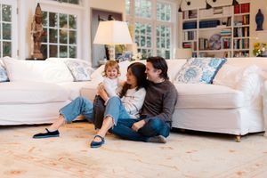 A family sits together on the floor in front of a white couch in a bright, cozy living room with built-in bookshelves.