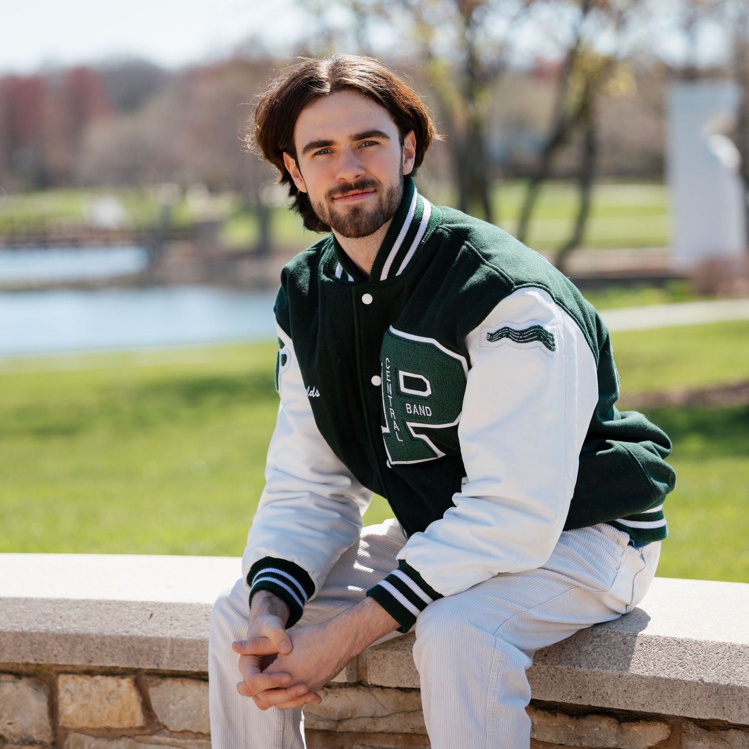Athletic person in green and white varsity jacket sits on stone wall near water.
