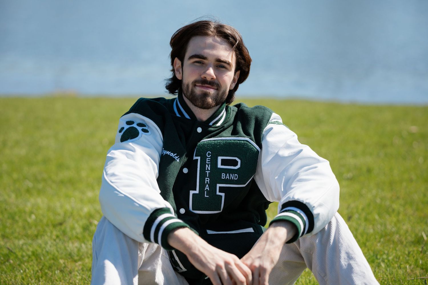Athletic varsity letterman in a green and white jacket sits on grass field.