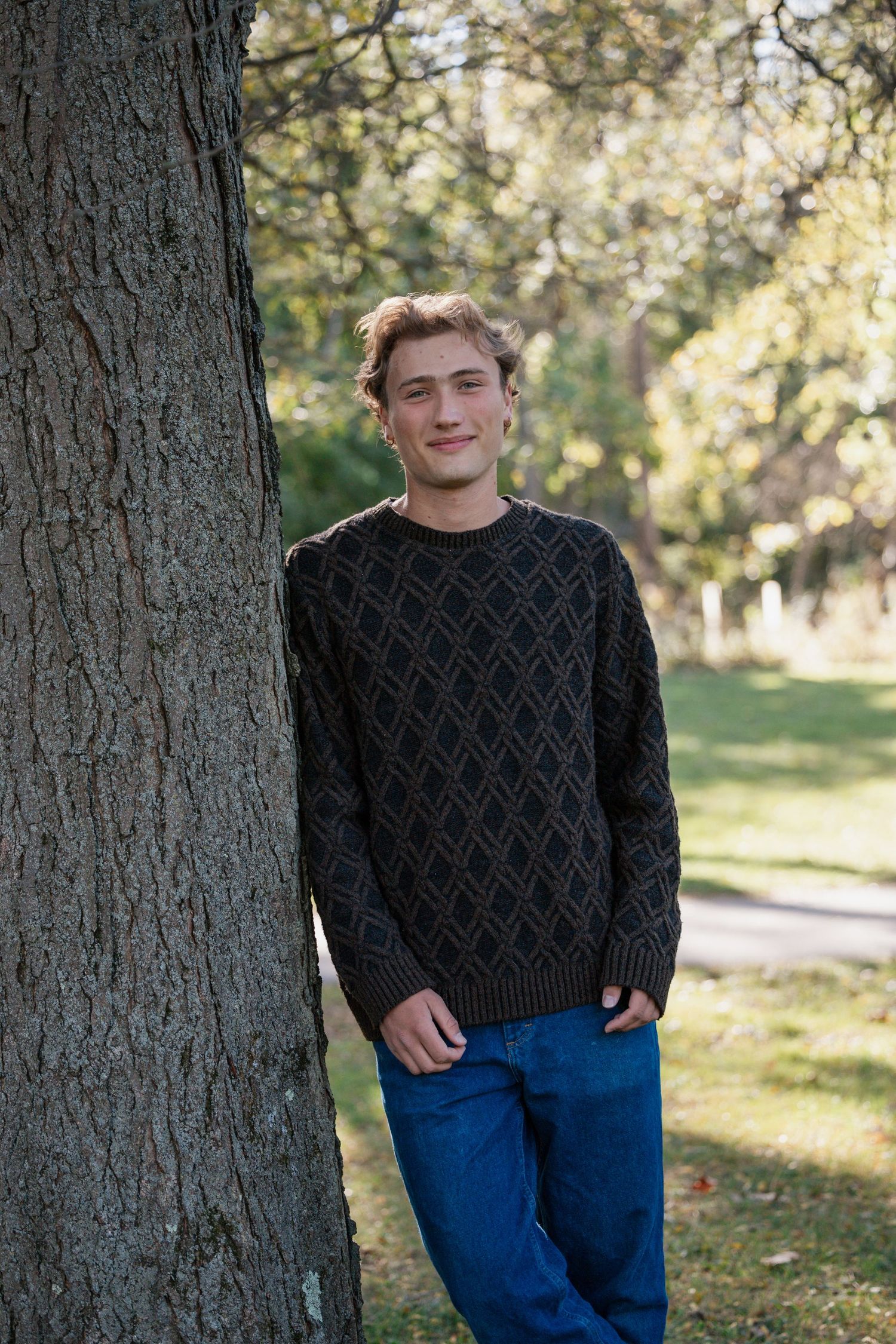 Young man in black sweater standing against a tree in a park setting.