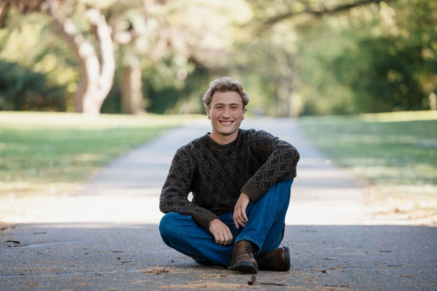 Person in dark sweater sitting cross-legged on a path in a leafy park.