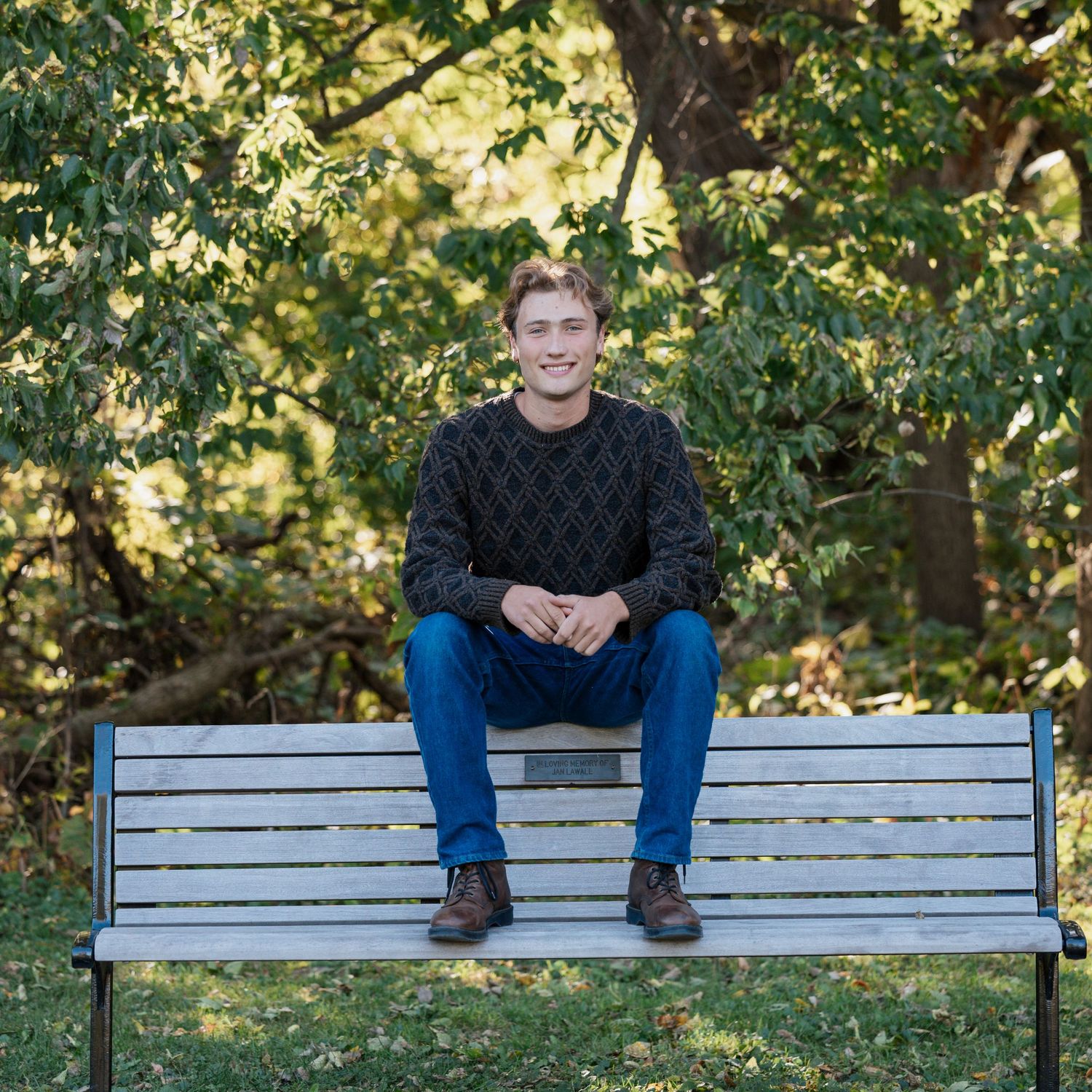 Person in cable knit sweater sitting on a white park bench surrounded by trees.