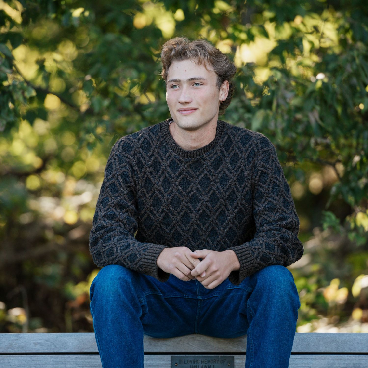 Person in a patterned sweater sits casually on a bench outdoors with green foliage in the background.