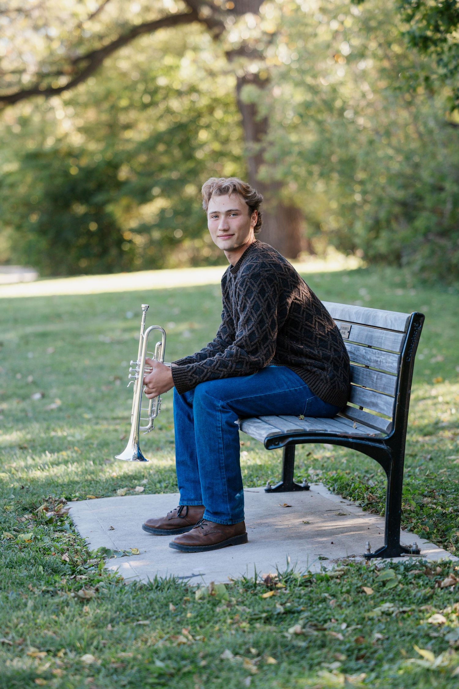 Musician in sweater sitting on park bench holding a trumpet among autumn trees.