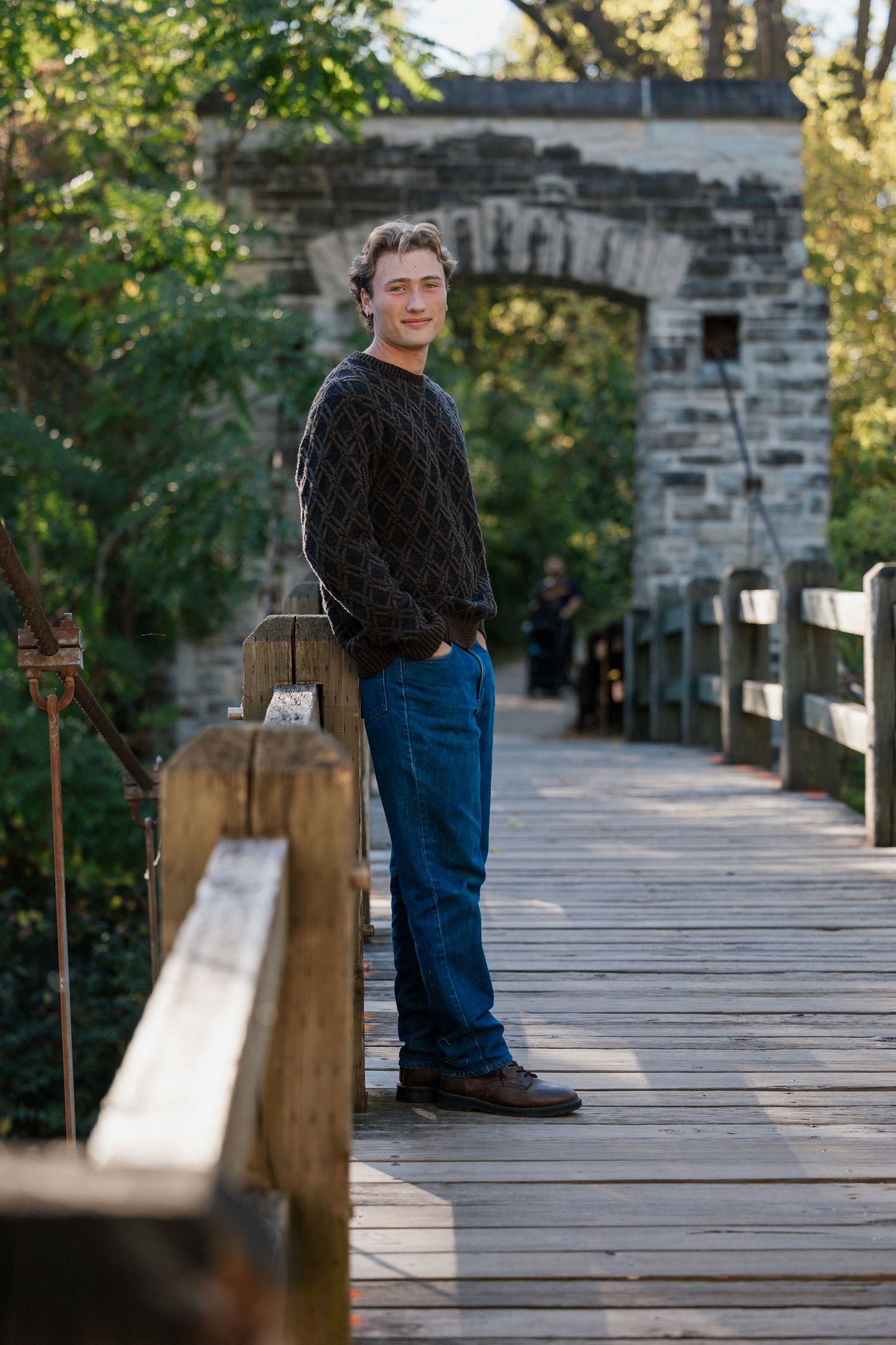 Person wearing a dark knit sweater and jeans stands on a wooden bridge with stone archway in a park setting.